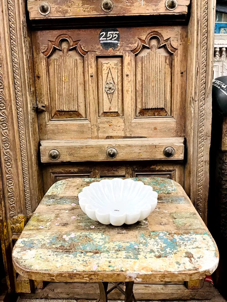May include: A white, scalloped bowl with a flower-like design rests on a distressed wooden table. The table has a multi-coloured, weathered appearance. The background features a vintage wooden door with decorative carvings and the number 255.