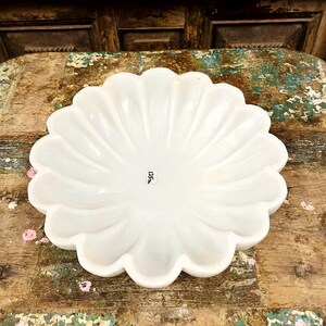 May include: A white, scalloped-edge decorative bowl. The bowl has a flower-like design with curved ridges radiating from the centre. The bowl is on a distressed wooden surface. The text "125" is visible on the bottom of the bowl.