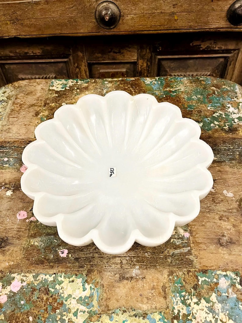 May include: A white, scalloped-edge decorative bowl. The bowl has a flower-like design with curved ridges radiating from the centre. The bowl is on a distressed wooden surface. The text "125" is visible on the bottom of the bowl.