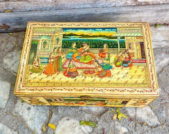 May include: A wooden box featuring a hand-painted scene of people in traditional attire. The box is adorned with a gold trim and elaborate floral patterns. The scene depicts a group of people gathered in a garden setting, with a palace in the background.