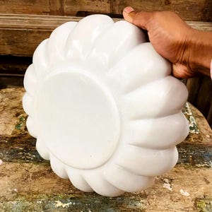 May include: A white, flower-shaped decorative bowl. The bowl has a smooth, glossy finish and a rounded base. The bowl is held by a hand, and the background is a wooden surface.