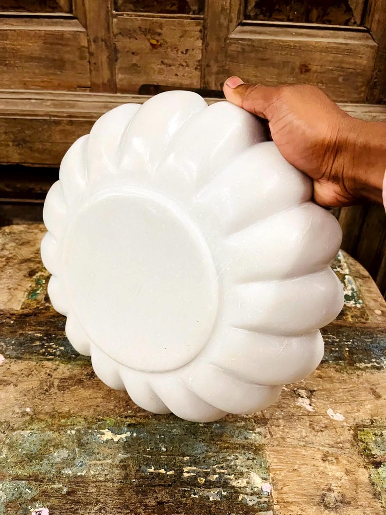 May include: A white, flower-shaped decorative bowl. The bowl has a smooth, glossy finish and a rounded base. The bowl is held by a hand, and the background is a wooden surface.