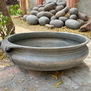 May include: A large, round, gray metal bowl with a decorative rim. The bowl is sitting on a stone surface.