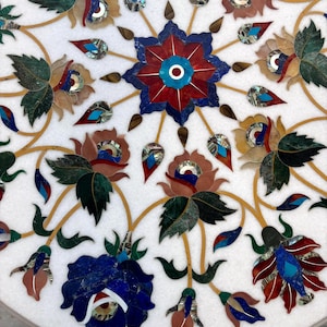 May include: Close-up of a white marble surface inlaid with a floral design. The design features a central starburst with red and blue petals, surrounded by various flowers and leaves in shades of green, brown, and blue. The details are accented with gold veins.