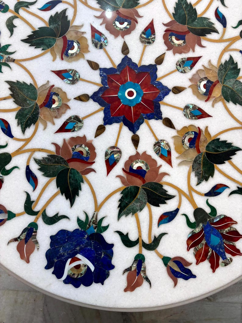 May include: Close-up of a white marble surface inlaid with a floral design. The design features a central starburst with red and blue petals, surrounded by various flowers and leaves in shades of green, brown, and blue. The details are accented with gold veins.