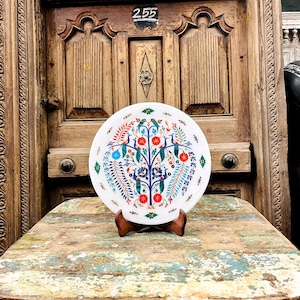May include: A white decorative plate with a colorful floral and peacock design. The plate is displayed on a wooden stand, set against a rustic wooden door background. The plate's design includes red, blue, and green accents.