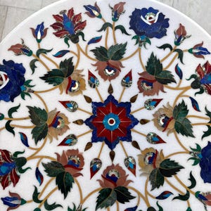 May include: A white marble table top with an inlaid floral design. The design features red, blue, green, and brown flowers with gold-colored stems and leaves. The flowers are arranged in a symmetrical pattern.