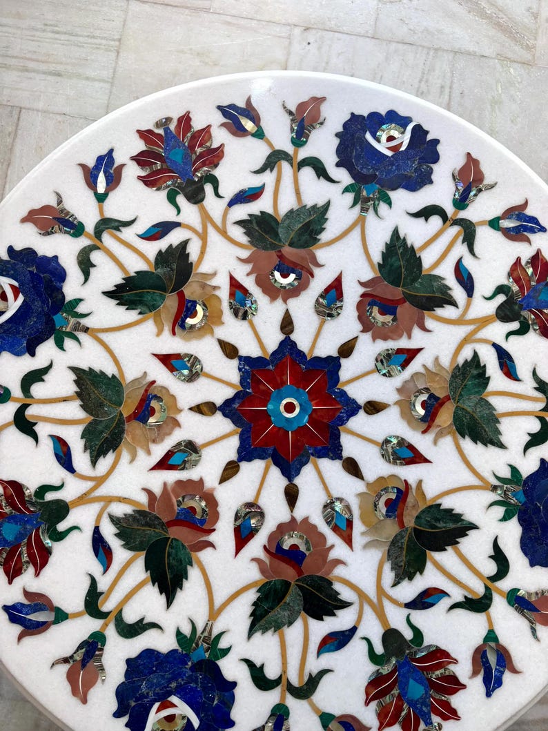 May include: A white marble table top with an inlaid floral design. The design features red, blue, green, and brown flowers with gold-colored stems and leaves. The flowers are arranged in a symmetrical pattern.