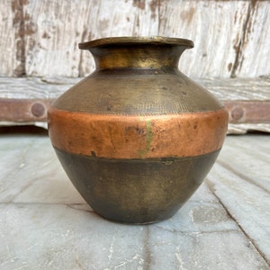 May include: A small, round, brass vase with a copper band around the middle. The vase has a dark brown patina and is sitting on a light gray surface.