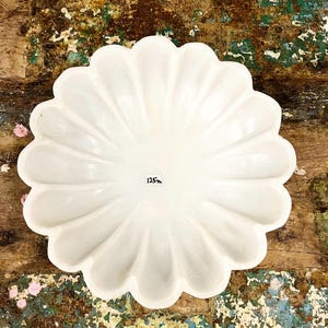 May include: A white, scalloped-edge serving dish. The dish has a flower-like shape with a series of curved petals. The number "125" is printed on the dish. The dish is set on a distressed wooden surface.