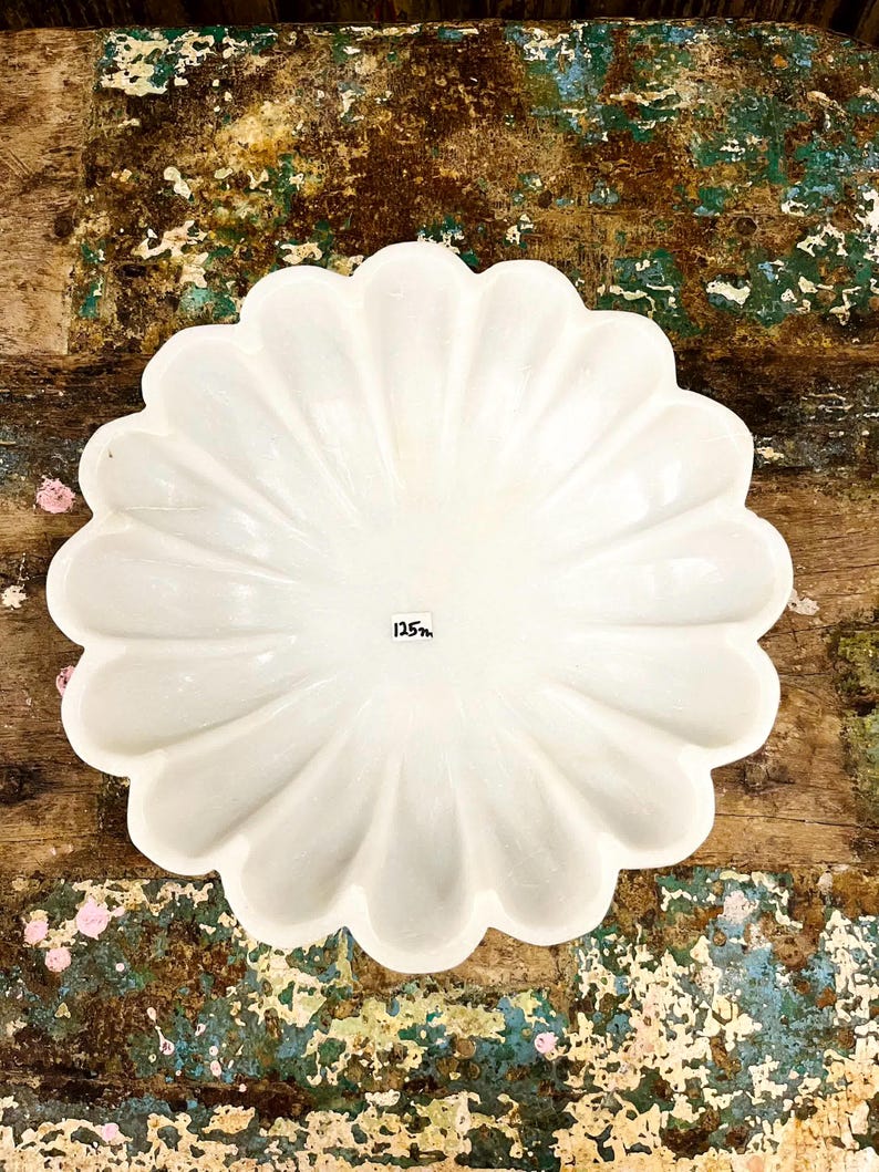 May include: A white, scalloped-edge serving dish. The dish has a flower-like shape with a series of curved petals. The number "125" is printed on the dish. The dish is set on a distressed wooden surface.