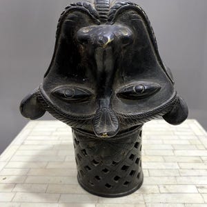 Può includere: Una scultura in metallo nero di un viso con occhi grandi e un design dettagliato. La scultura è montata su una base cilindrica con un motivo a traliccio.