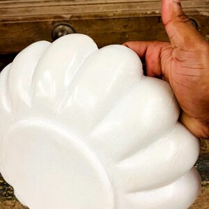 May include: A white, flower-shaped decorative object with a smooth, glossy finish. The object has a series of rounded petals radiating from a central point. The image shows the object being held by a hand.