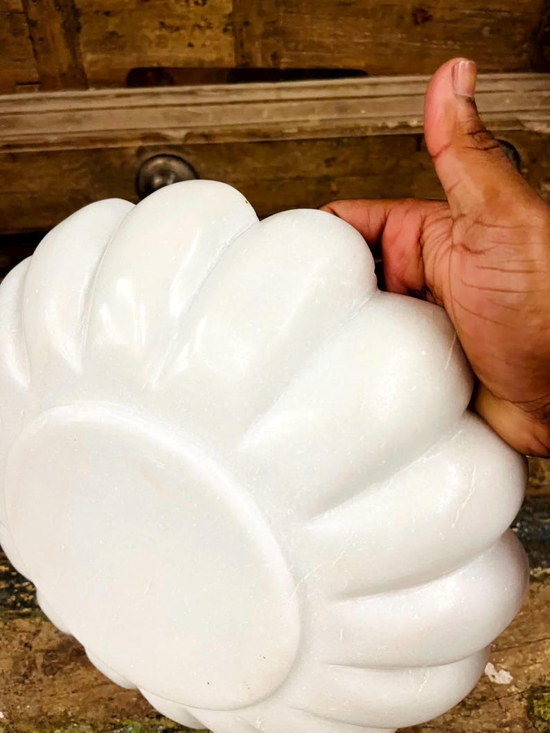 May include: A white, flower-shaped decorative object with a smooth, glossy finish. The object has a series of rounded petals radiating from a central point. The image shows the object being held by a hand.