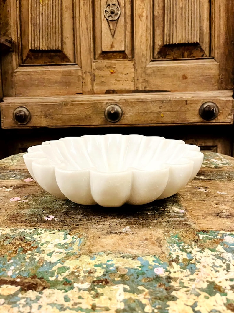 May include: A white marble bowl with a scalloped edge, resembling a flower. The bowl sits on a distressed wooden surface. The background features a wooden door. The bowl's design has rounded petals.