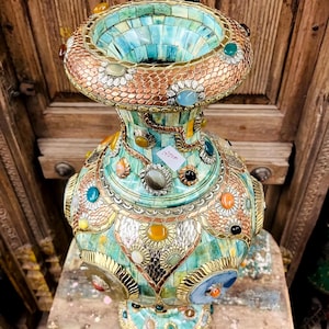 Puede incluir: Un jarrón decorativo con un diseño de mosaico en tonos turquesa, dorado y cobre. El jarrón está adornado con piedras preciosas de colores y trabajos de metal intrincados. Tiene una parte superior ancha y acampanada y un cuerpo bulboso, que descansa sobre una base.