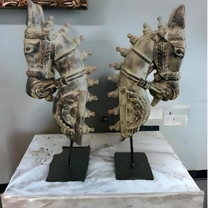 Puede incluir: Dos esculturas antiguas de madera tallada con forma de cabeza de caballo con intrincados detalles, montadas sobre soportes de metal negro. Las esculturas son de color gris claro y tienen un acabado desgastado.