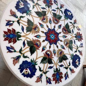 May include: Round white marble table with intricate floral inlay design. The design features blue, red, green, and brown floral patterns. The table's surface is polished and smooth, showcasing the detailed craftsmanship.
