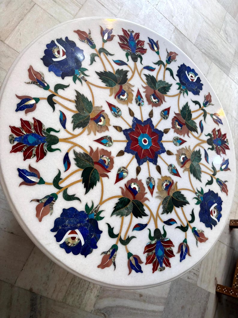 May include: Round white marble table with intricate floral inlay design. The design features blue, red, green, and brown floral patterns. The table's surface is polished and smooth, showcasing the detailed craftsmanship.