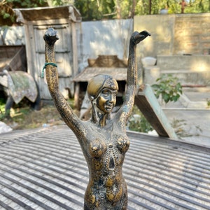 May include: A gold-toned metal sculpture of a woman with her arms raised above her head. The sculpture is detailed with a textured surface and intricate patterns.