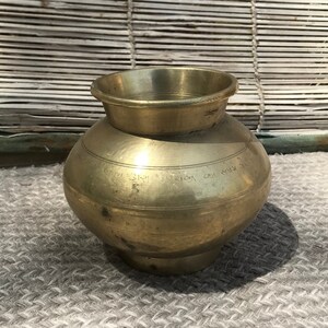 May include: A small, round, brass pot with a wide mouth and a narrow base. The pot has a smooth, shiny surface and is inscribed with text in an unknown language.