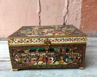 May include: A small wooden box with a hinged lid, decorated with intricate hand-painted scenes of people and animals in a procession. The box is painted in vibrant colours, including gold, red, blue, and green. The lid is closed, and the box is sitting on a white marble surface.