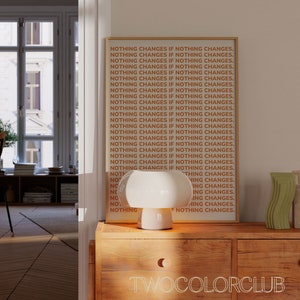 May include: A framed print with the text "NOTHING CHANGES IF NOTHING CHANGES." repeated multiple times in orange and white on a beige background. The print is hanging on a wall in a room with a white lamp and decorative objects on a wooden table.