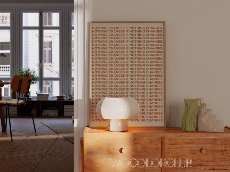 May include: A framed print with the text "NOTHING CHANGES IF NOTHING CHANGES." repeated multiple times in orange and white on a beige background. The print is hanging on a wall in a room with a white lamp and decorative objects on a wooden table.