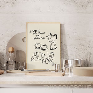 May include: A framed print with the text "Sundays are made for breakfast" and a drawing of a coffee pot, two cups of coffee, and two croissants. The print is on a white countertop with a wooden chopping board, a glass bottle, and other kitchen items.