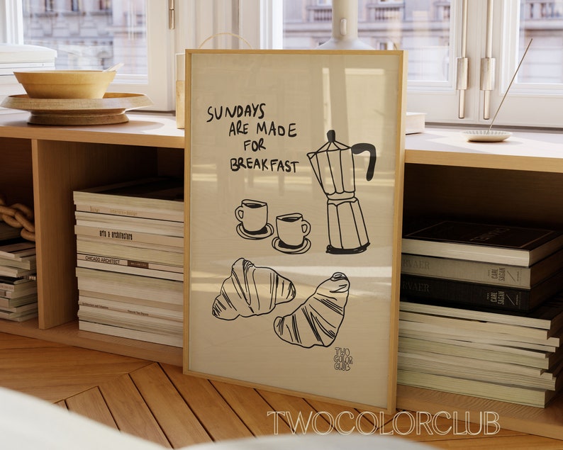 May include: A framed print with the text "Sundays are made for breakfast" and a drawing of a coffee pot, two coffee cups, and two croissants. The print is on a light brown background and has a wooden frame.