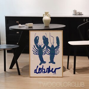 以下が含まれることがあります： 2匹の青いロブスターのイラストと、青いスクリプトで「lobster」という言葉が描かれた額入りのアートプリント。 黒いテーブル、椅子、白い壁のある部屋にプリントが飾られています。