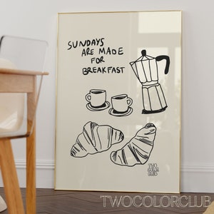 May include: A white framed print with black text that reads "Sundays are made for breakfast". The print also features a black and white illustration of a coffee pot, two coffee cups, and two croissants.