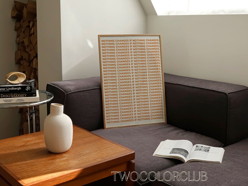 May include: A framed print with the text "NOTHING CHANGES IF NOTHING CHANGES" repeated in a light orange color on a white background. The print is hanging on a wall in a living room setting.