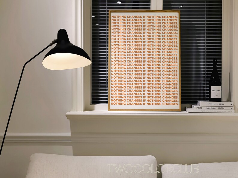 May include: A framed print with the text "NOTHING CHANGES IF NOTHING CHANGES." repeated in a light orange color on a white background. The print is hanging on a white wall above a window with white blinds.