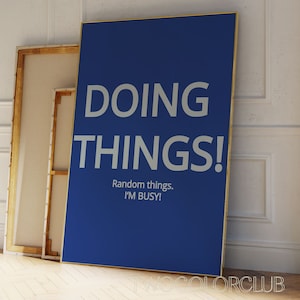 May include: A blue poster with white text that reads "DOING THINGS! Random things. I'M BUSY!" The poster is framed in a gold frame and is leaning against a white wall.