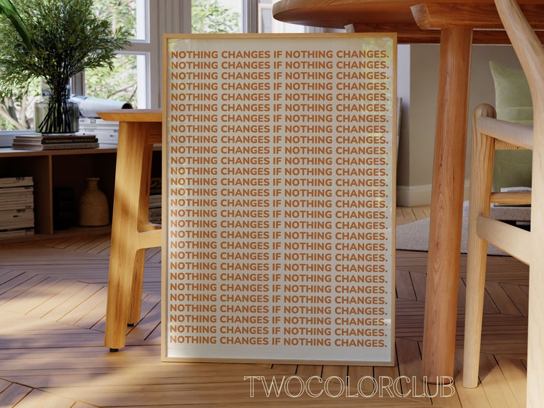 May include: A framed print with the text "NOTHING CHANGES IF NOTHING CHANGES." repeated 28 times in a light orange color on a white background. The frame is a light wood color. The text "TWOCOLORCLUB" is printed in black at the bottom of the print.