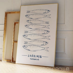 May include: A white canvas print with a gold frame featuring a minimalist design of nine blue sardines in a row. The text "SARDINES" and "TWOCOLORCLUB" is printed in blue below the sardines.