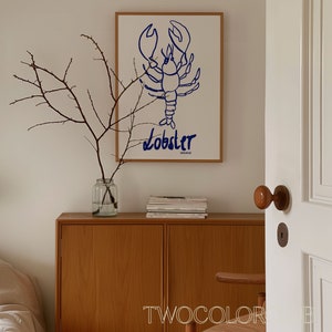 K&ouml;nnte beinhalten: Ein wei&szlig;er gerahmter Druck mit einer blauen Linienzeichnung eines Hummers und dem Wort "Lobster" darunter. Der Druck h&auml;ngt an einer wei&szlig;en Wand &uuml;ber einem Holzschr&auml;nkchen mit einer Vase mit Zweigen und einem Stapel B&uuml;chern.