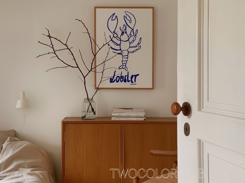 K&ouml;nnte beinhalten: Ein wei&szlig;er gerahmter Druck mit einer blauen Linienzeichnung eines Hummers und dem Wort "Lobster" darunter. Der Druck h&auml;ngt an einer wei&szlig;en Wand &uuml;ber einem Holzschr&auml;nkchen mit einer Vase mit Zweigen und einem Stapel B&uuml;chern.