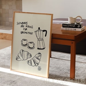May include: A framed print with a black and white illustration of a coffee pot, two coffee cups, and croissants. The text on the print reads "Sundays are made for breakfast".