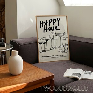 May include: A framed black and white illustration of glasses with the text "HAPPY HOUR" and "WEEK NIGHTS 6 PM - 11:30 PM SHOULD WE GET ANOTHER DRINK?"  The illustration is on a white background with a wooden frame. The frame is sitting on a gray couch with a book on the couch.
