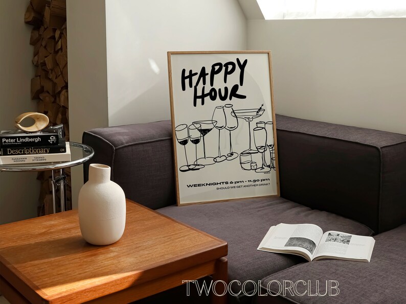 May include: A framed black and white illustration of glasses with the text "HAPPY HOUR" and "WEEK NIGHTS 6 PM - 11:30 PM SHOULD WE GET ANOTHER DRINK?"  The illustration is on a white background with a wooden frame. The frame is sitting on a gray couch with a book on the couch.