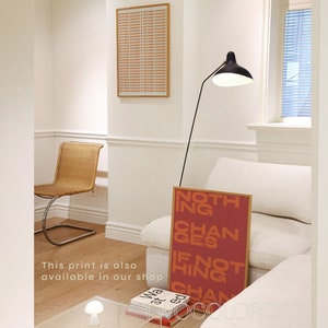 May include: A red and orange print with the text "Nothing Changes If Nothing Changes" hanging on a white wall in a living room. The print is framed in a light wood frame.