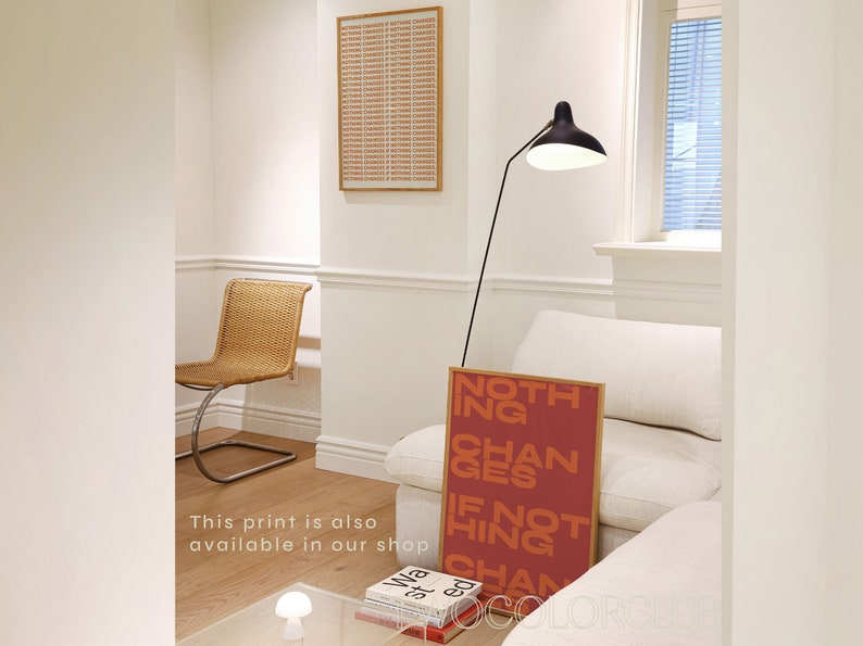 May include: A red and orange print with the text "Nothing Changes If Nothing Changes" hanging on a white wall in a living room. The print is framed in a light wood frame.