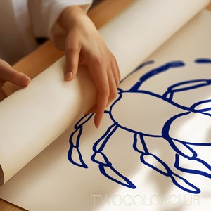 K&ouml;nnte beinhalten: Eine Person rollt ein wei&szlig;es Blatt Papier auf, auf dem eine blaue Tintenzeichnung eines Krebses zu sehen ist. Die Zeichnung ist einfach und stilisiert, die Beine und der K&ouml;rper des Krebses sind mit dicken blauen Linien umrandet. Der Text "TWOCOLORCLUB" ist unten auf dem Blatt gedruckt.