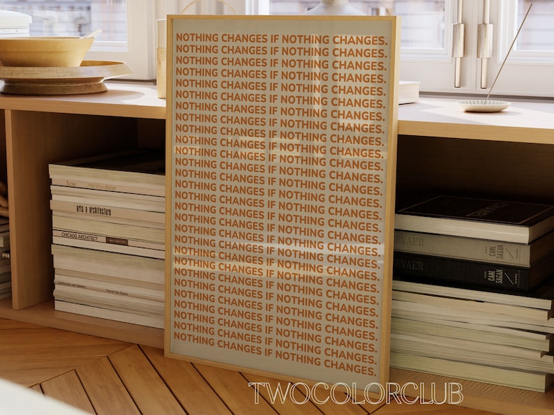 May include: A framed print with the text "NOTHING CHANGES IF NOTHING CHANGES." repeated 28 times. The text is in orange on a white background. The frame is light brown wood. The print is on a shelf with books.