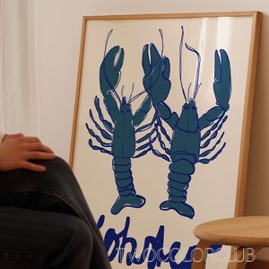以下が含まれることがあります： クリーム色の背景に、2匹の様式化された青いロブスターが描かれた額入りのアートプリント。「Lobster」の文字が青色で下に印刷されています。プリントは明るい木製のフレームに入っています。