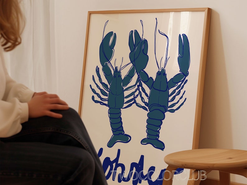 以下が含まれることがあります： クリーム色の背景に、2匹の様式化された青いロブスターが描かれた額入りのアートプリント。「Lobster」の文字が青色で下に印刷されています。プリントは明るい木製のフレームに入っています。