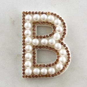 Peut inclure: Une lettre "B" décorative blanche, ornée de fausses perles et entourée de petits strass couleur bronze. La lettre est posée sur une surface en marbre blanc avec des accents dorés et un fond en velours rose.