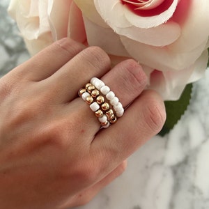 May include: A set of three white beaded rings with gold accents. The rings are stacked on a finger.